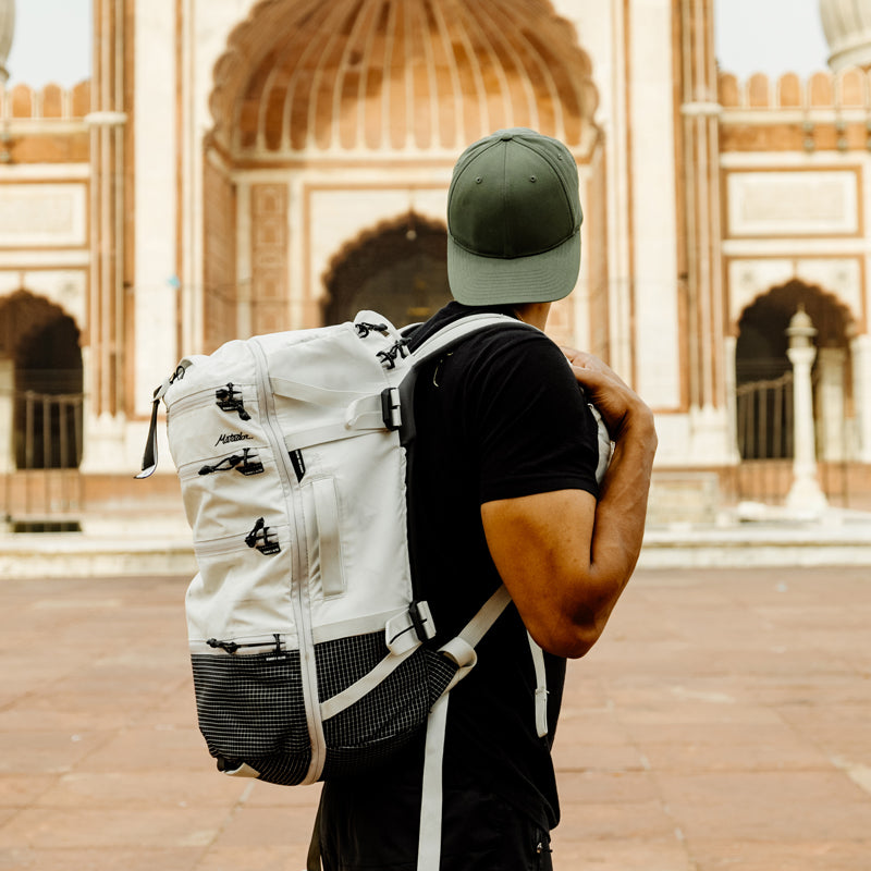 Man in green backwards hat in front of Indian temple, wearing white SEG28 backpack